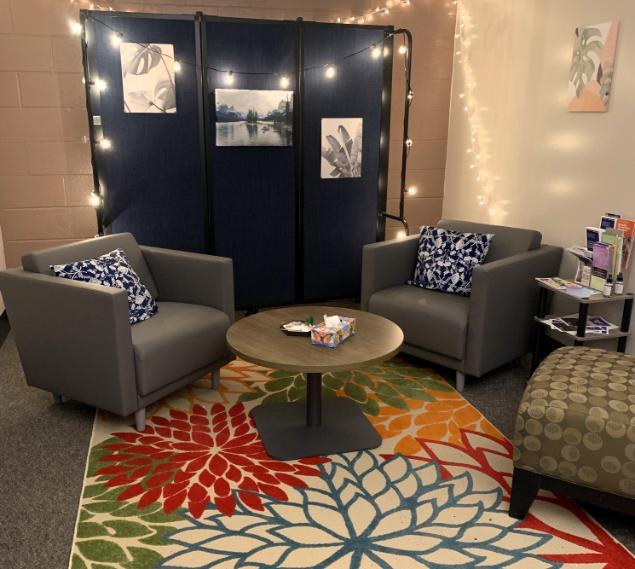 An image of the Oasis/Haven location - two gray comfortable lounge chairs with a table in the middle and a colorful carpet. There are string lights behind and a small table with informational materials on the right.